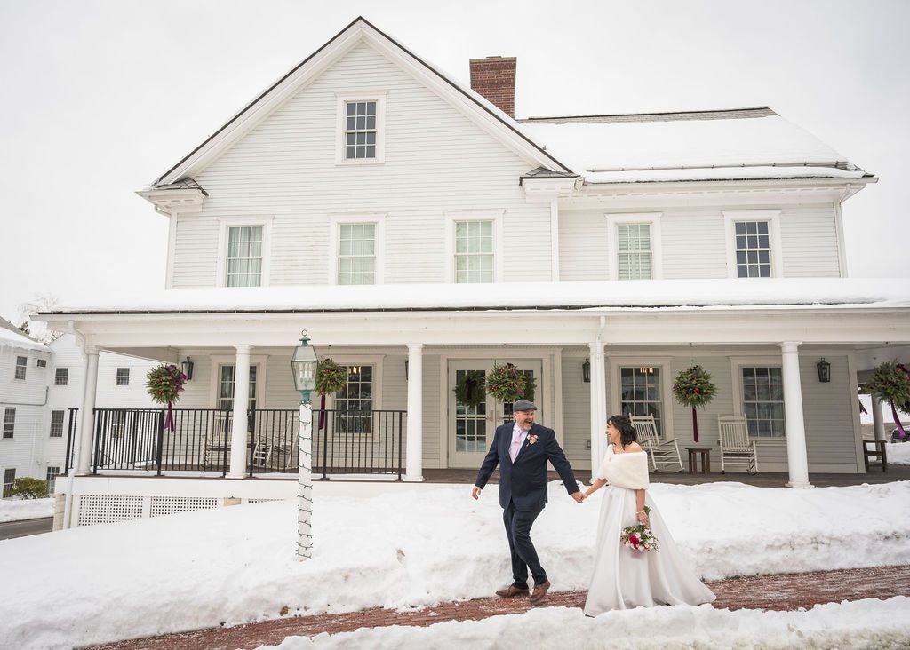 Wedding couple poses in front of a classic white farmhouse with wrap-around porch in winter.