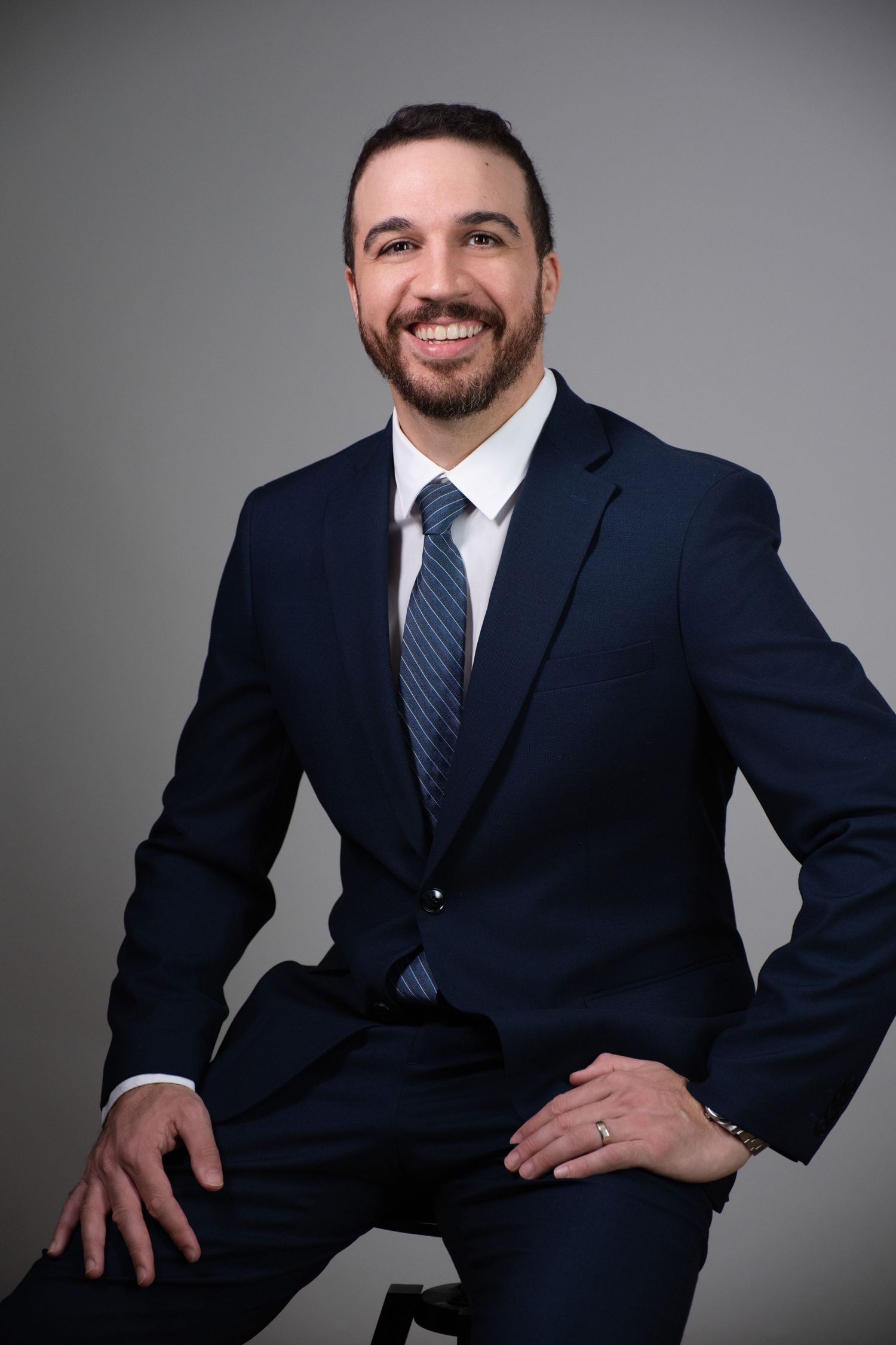 Professional headshot of someone in a navy suit and blue striped tie smiling against a gray background.