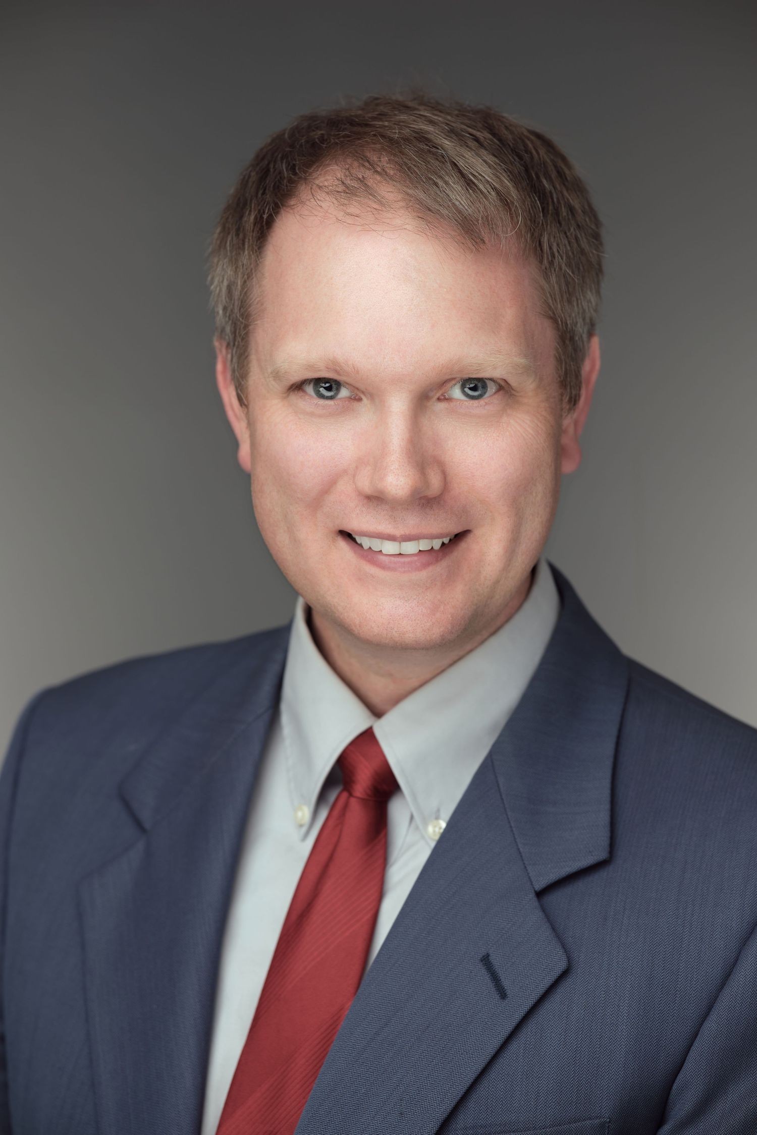 Corporate headshot of a professional in a navy suit against a gray background