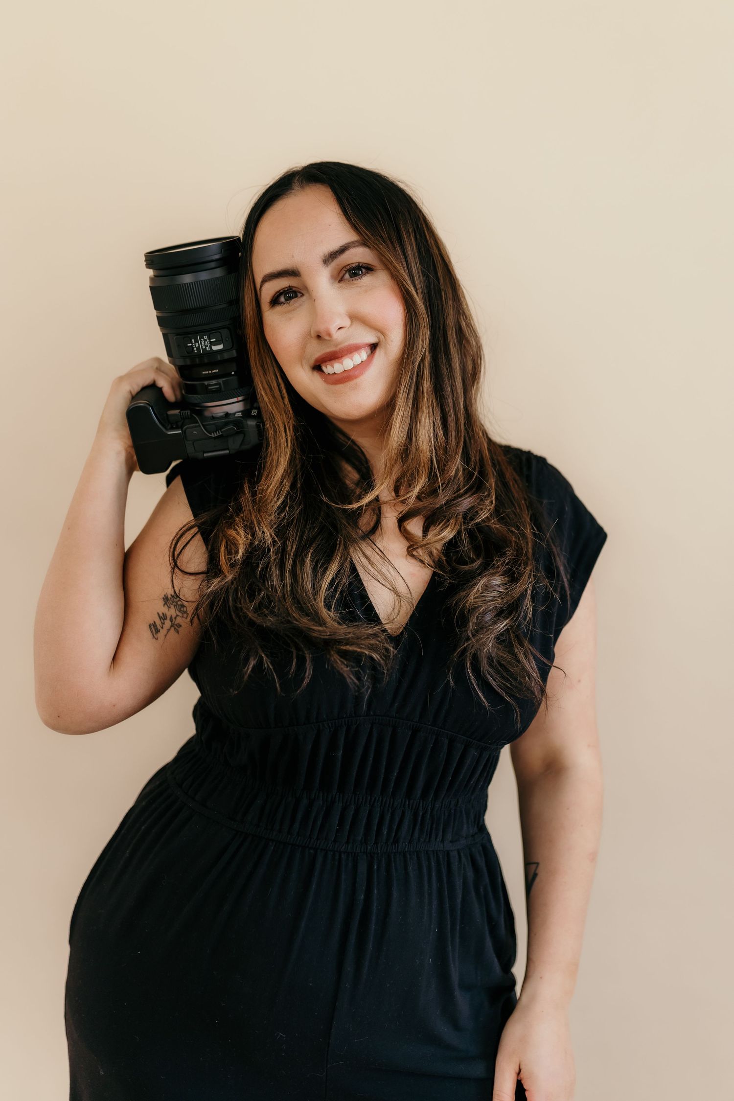 A photographer in casual wear sits cross-legged with camera against beige background.