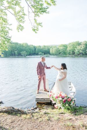 Wedding couple stands on a dock over a lake with vibrant floral decorations.