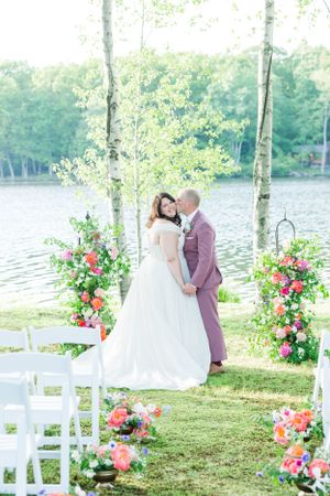 Wedding ceremony setup by lake with white chairs and pink floral arrangements framing the aisle.
