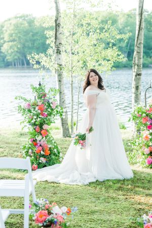 A bride in white stands between birch trees and floral arrangements by a lakeside ceremony site.