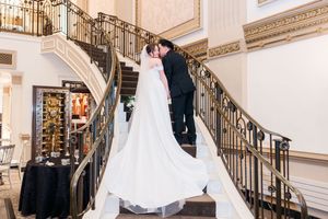 Bride and groom pose together near grand staircase decorated with white floral arrangements in opulent venue.