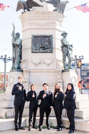 A wedding party poses playfully in front of a historic monument with American flags and stone statues.