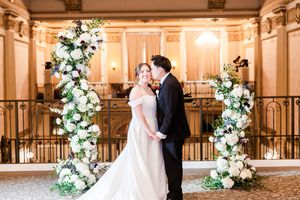 Elegant wedding couple shares romantic moment in luxurious ballroom with ornate staircase and floral arrangements.