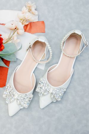 Elegant white bridal shoes with crystal embellishments displayed with delicate pink flowers on a gray background.