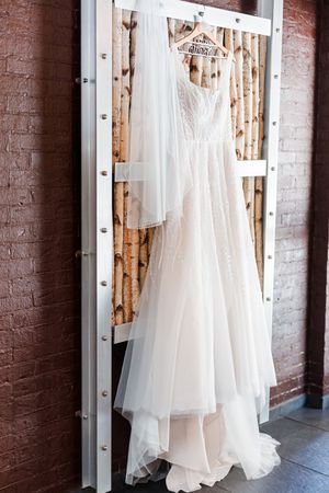 An elegant white lace wedding dress hangs on display against a rustic brick wall with birch branches in the background.