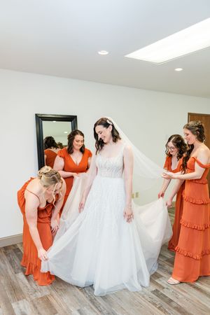 A wedding party in a bridal suite with bridesmaids in coral orange dresses helping with the white wedding gown.