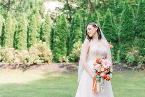 A bride in a flowing white gown holds a vibrant coral and white bouquet while standing in a lush garden setting.