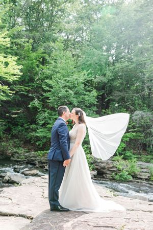 A couple in wedding attire embraces on a rocky outcrop near a flowing stream surrounded by lush green forest.