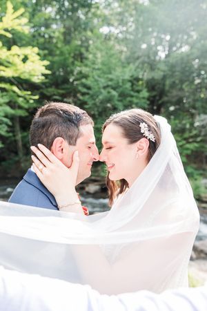 A romantic wedding moment captured outdoors with a flowing veil against a lush green forest backdrop.