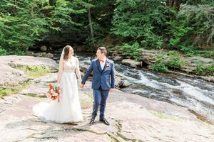 A couple in wedding attire stands together on rocky terrain beside a flowing stream in a lush forest setting.