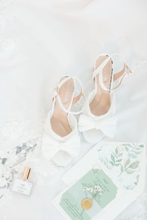 Elegant white bridal shoes with ankle straps styled with perfume and mint green invitation on marble surface.