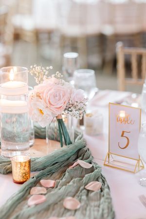 Wedding table decor features soft pink roses, sage green fabric, and gold table numbers with glowing candle votives.