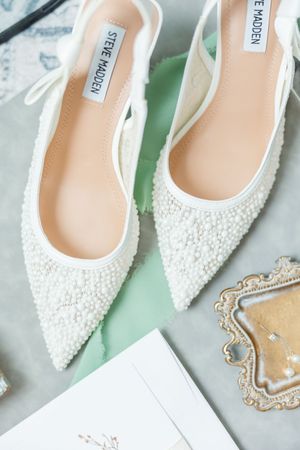 Elegant white pearl-encrusted bridal shoes with pointed toes displayed on a marble and mint green surface.