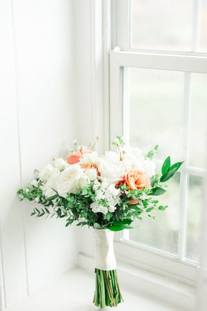 A delicate bridal bouquet with peach and white roses, greenery and foliage arranged by a bright window.