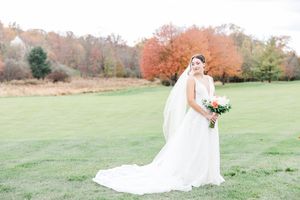 A bride in a flowing white gown stands in an autumn field with orange and red trees in the background.