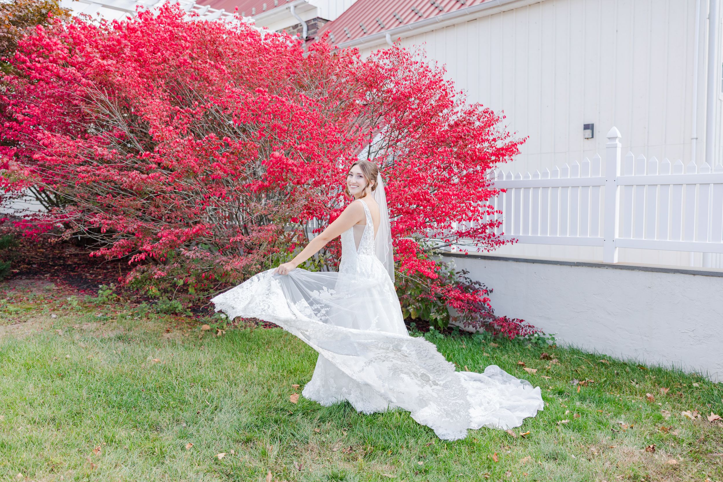 White wedding dress flowing in front of a white fence with vibrant red autumn foliage in the background.