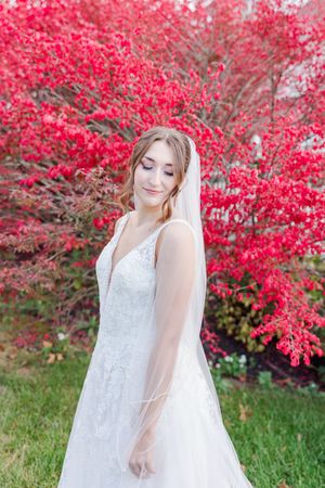 A bride in a white lace wedding dress poses gracefully against vibrant red autumn foliage in an outdoor setting.
