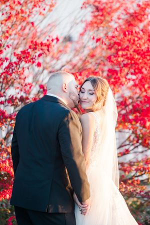 A couple shares an intimate moment in front of vibrant red autumn foliage while wearing wedding attire.