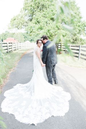 Elegant bride in lace wedding gown with dramatic train posing on paved path with wooden fence.