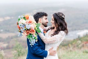 A couple in wedding attire embraces on a scenic mountain overlook with soft floral bouquet accents.