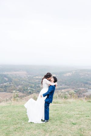 A couple in wedding attire embraces on a hilltop meadow with scenic mountain views in the background.
