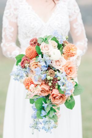 A beautiful wedding bouquet featuring coral roses, peach dahlias, and light blue delphinium flowers arranged elegantly.