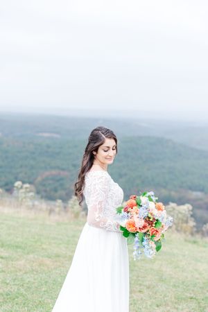 A bride in a white lace dress poses with a colorful bouquet against a misty mountain landscape backdrop.