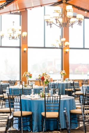 Wedding reception venue with round tables draped in dusty blue linens and dark wooden chiavari chairs.