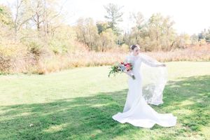 A flowing white wedding dress and veil catch the breeze in a sunlit autumn field.