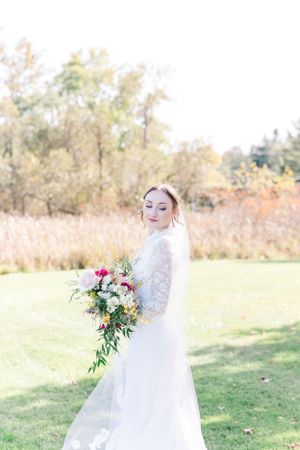 Bride in lace gown holds cascading bouquet while posing in sunlit autumn field.