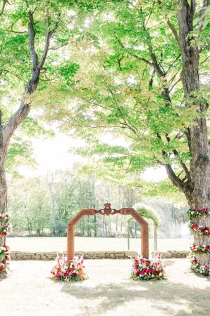 A romantic wedding arch decorated with pink flowers under a canopy of green trees.