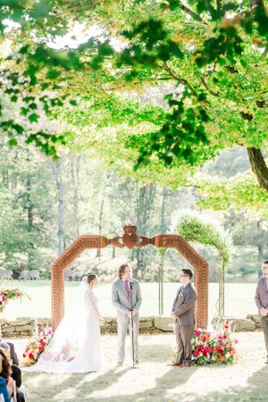 Outdoor wedding ceremony under rustic wooden arch decorated with flowers in a sunlit forest setting.