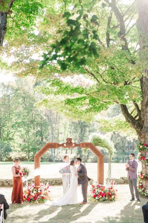 Sunlit wedding ceremony under wooden arch with red floral arrangements in forest setting.