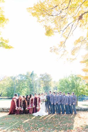 Wedding party in burgundy dresses and gray suits poses together under golden autumn foliage in an outdoor setting.