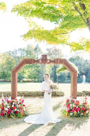 Bride poses under ornate wooden arch decorated with pink and red flowers in autumn setting.