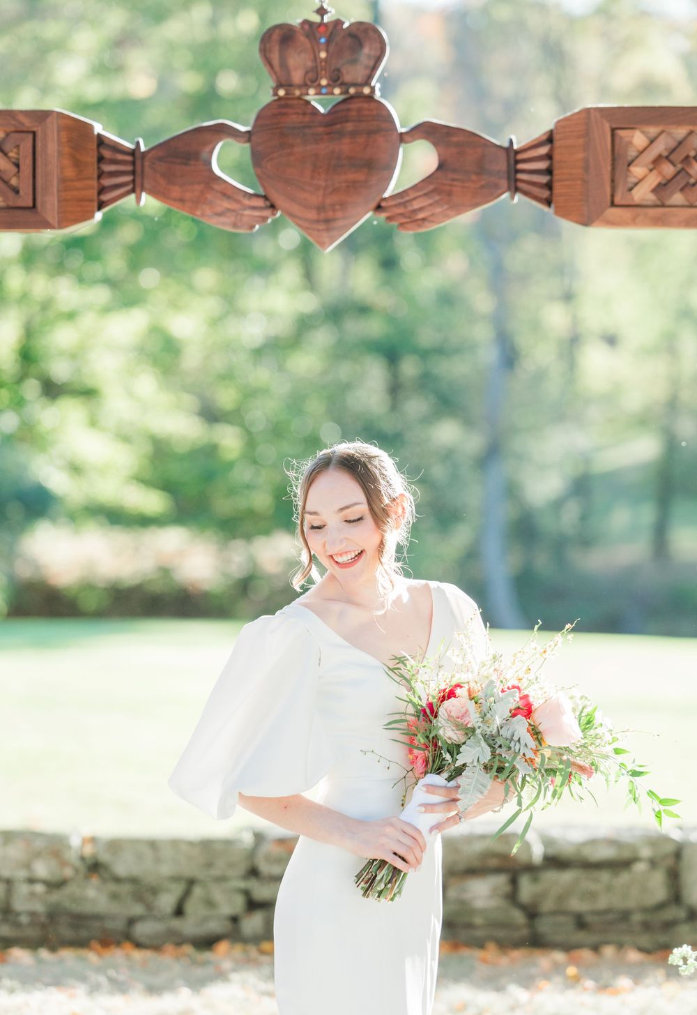 A bride poses beneath a wooden Claddagh symbol holding a delicate bouquet.