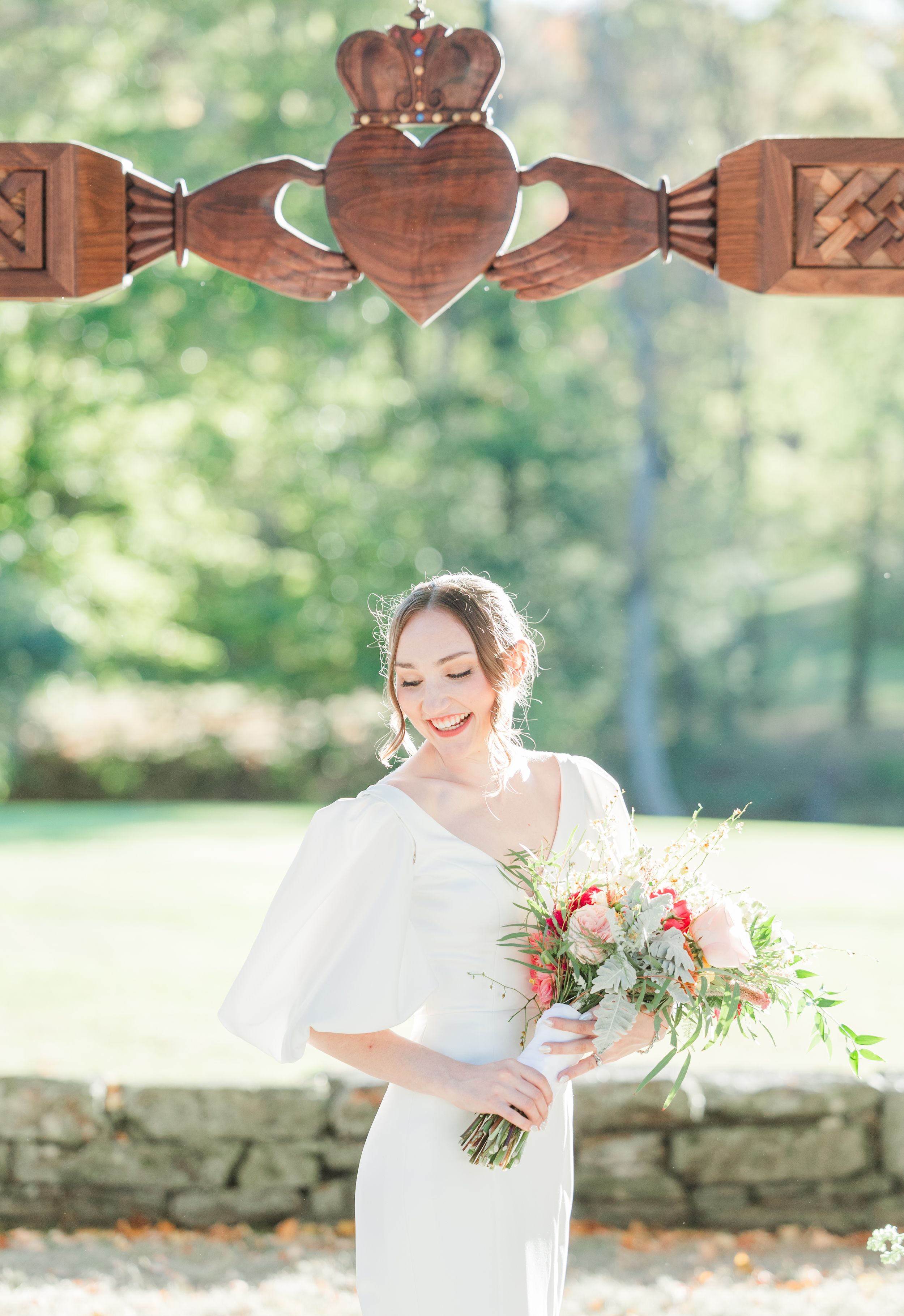 A bride poses beneath a wooden Claddagh symbol holding a delicate bouquet.