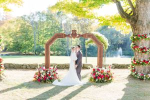 Couple stands under a wooden arch with red floral arrangements during autumn wedding.