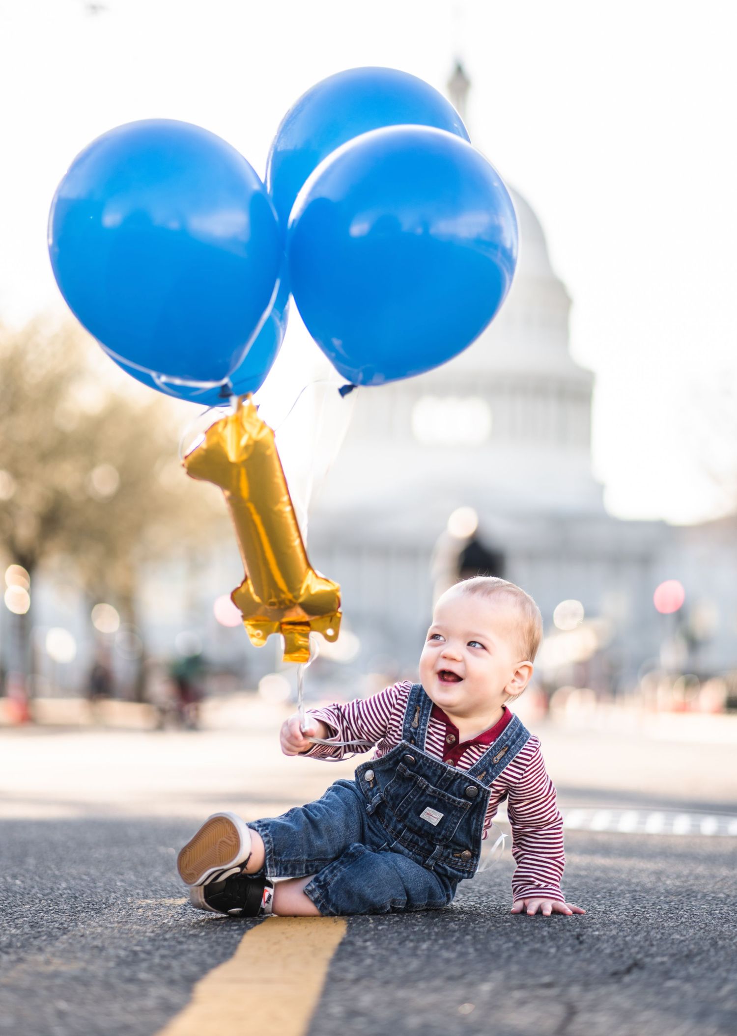 A baby in overalls lies on pavement while holding blue balloons with gold number 4 decorations.