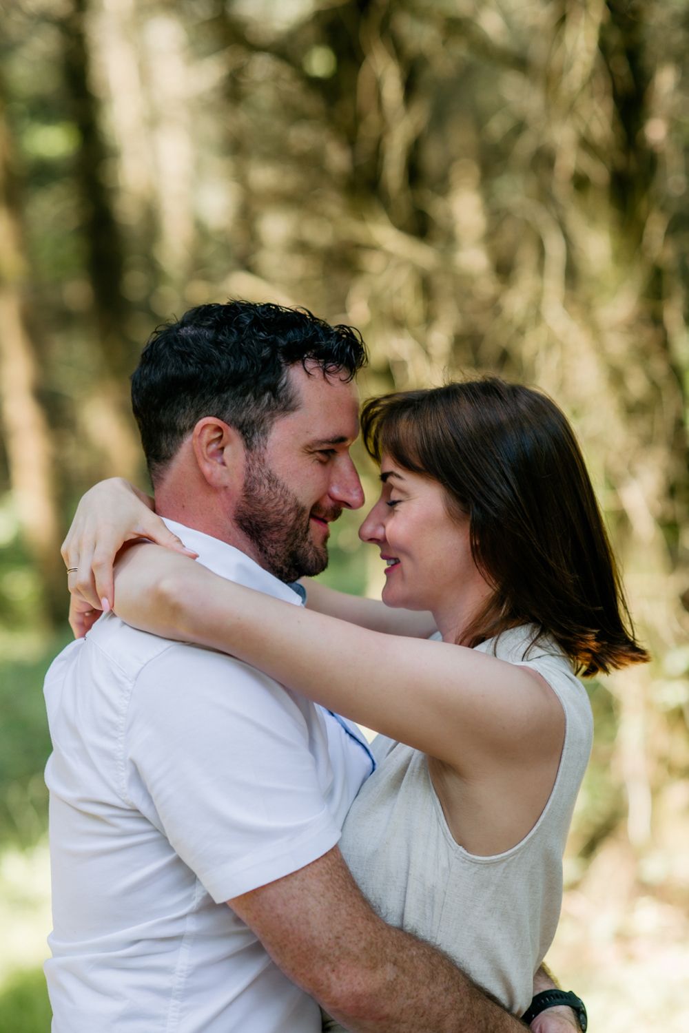 Romantic outdoor portrait of a couple embracing with foreheads touching in a sunlit forest, natural candid moment, soft background bokeh.