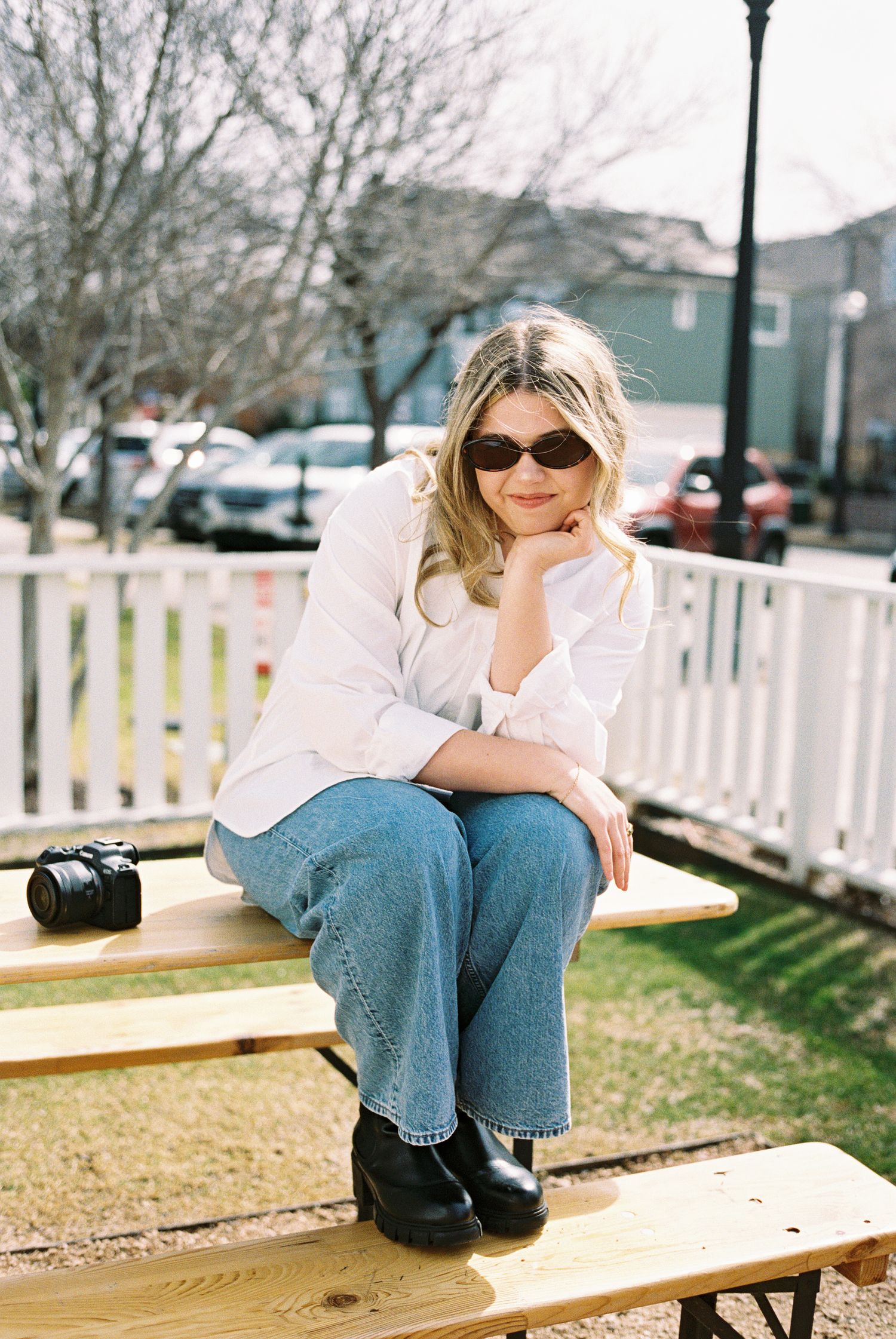 Blonde woman in white blazer and jeans sitting on a bench outdoors with a camera beside her.