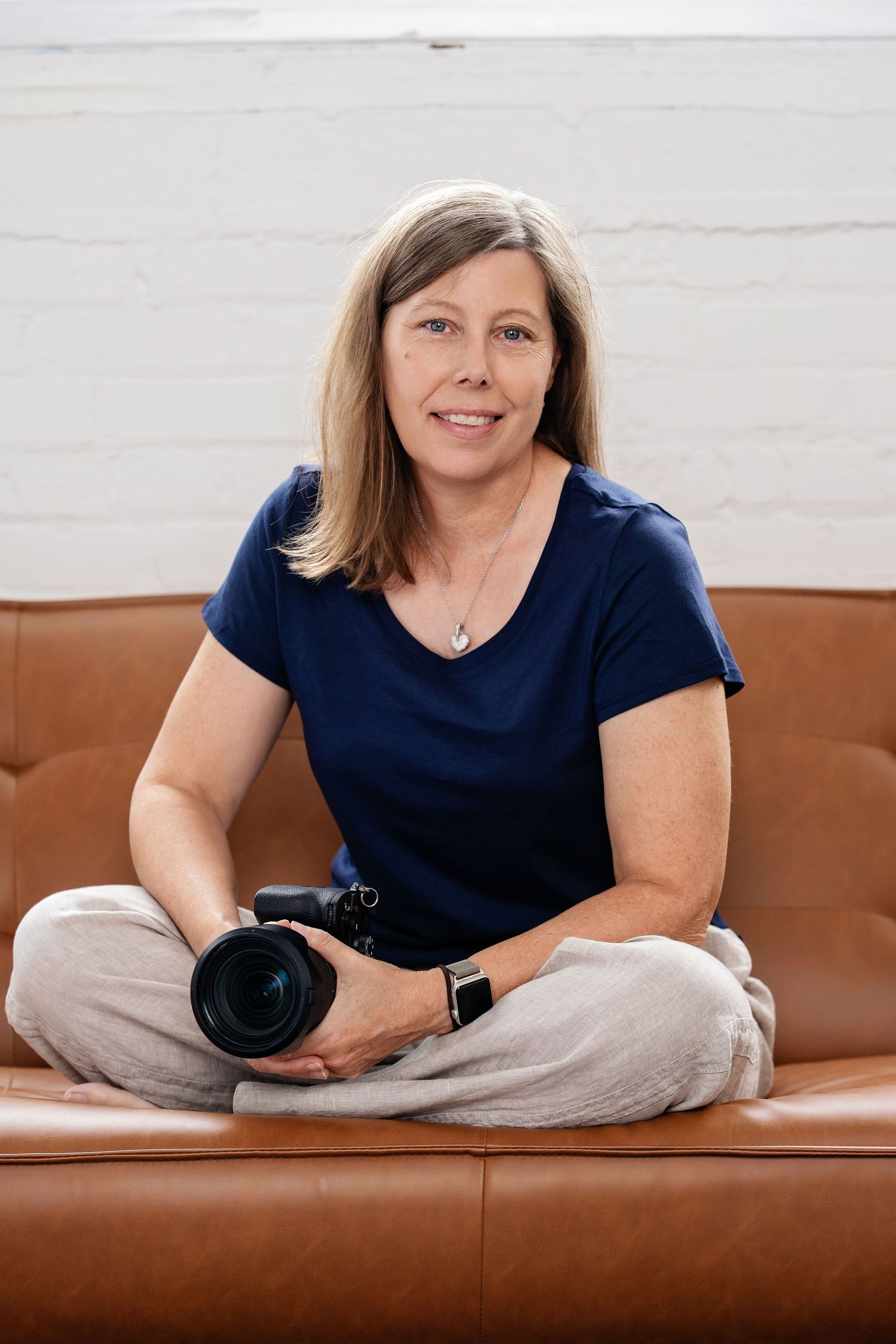 Milwaukee senior, family, headshot and event photographer Rachel Dickman sitting cross legged on a sofa smiling at camera holding camera.