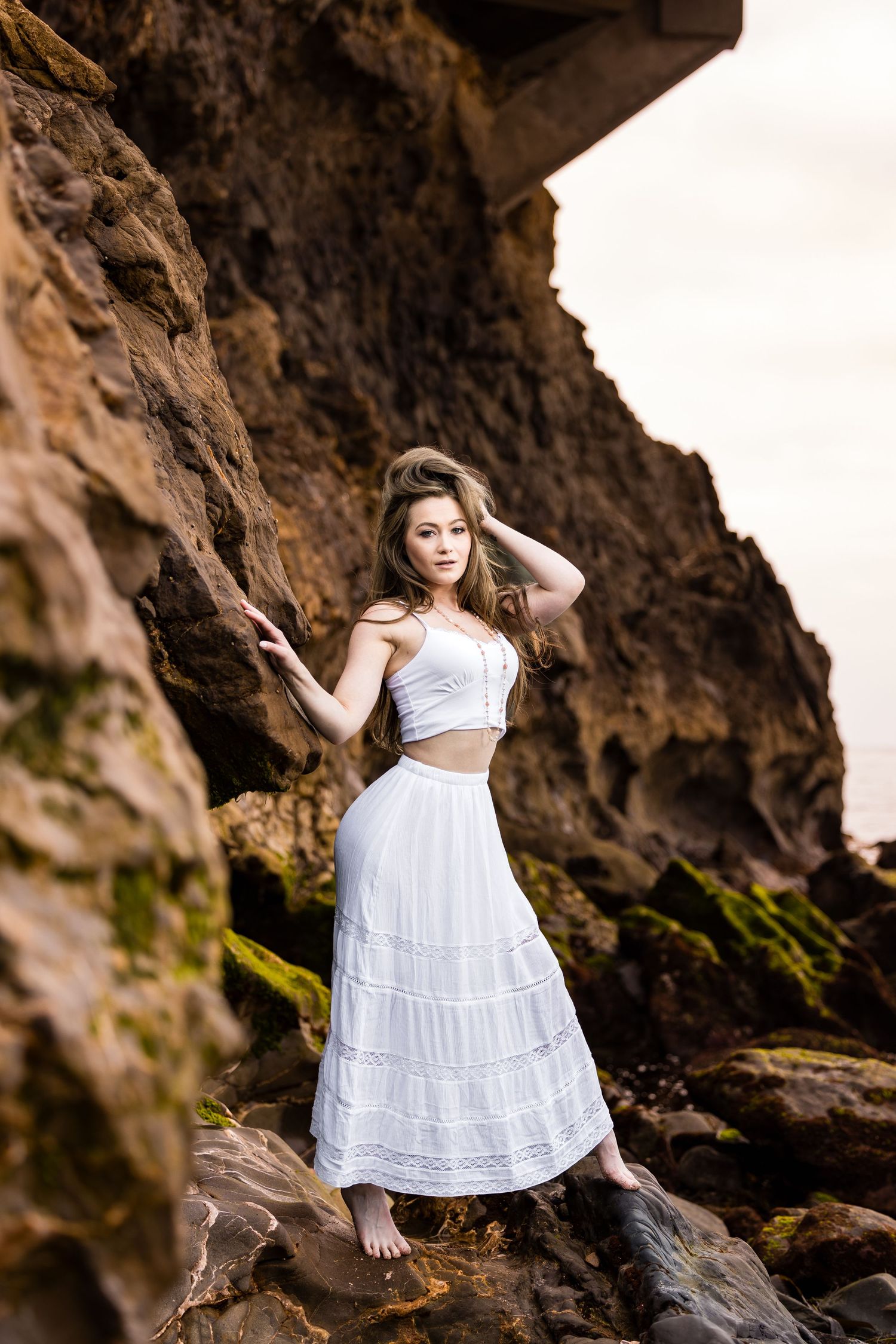 Woman in white two-piece outfit poses on rocky beach formation at sunset.