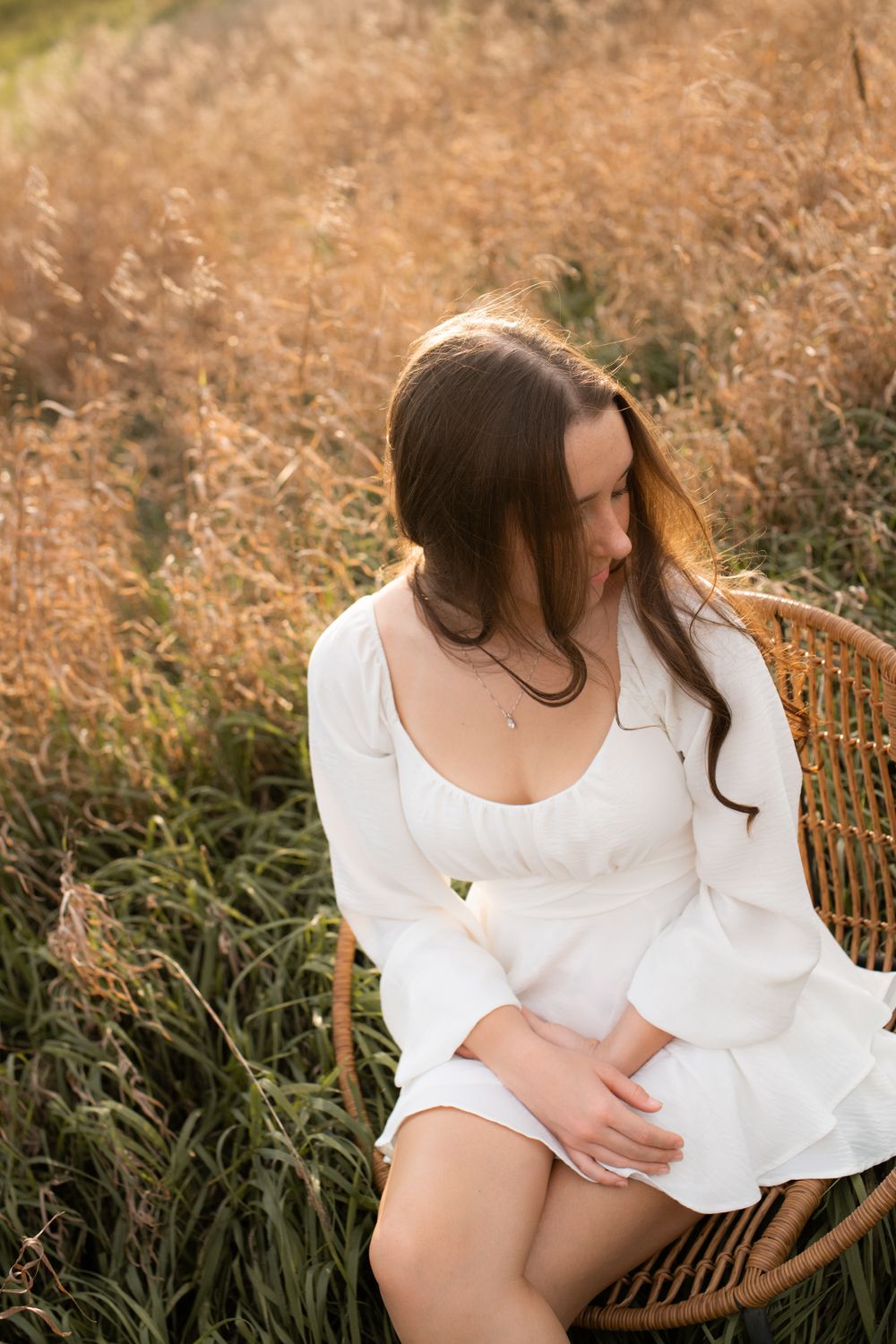 A high school senior in a white flowy dress poses gracefully in a golden field during a golden hour sunset senior photo session.