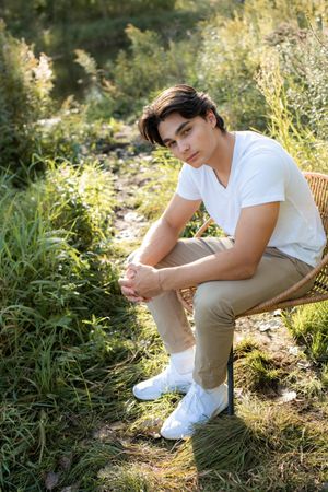 A high school senior in white shirt and khaki pants poses casually on grassy ground during a sunny summer day.
