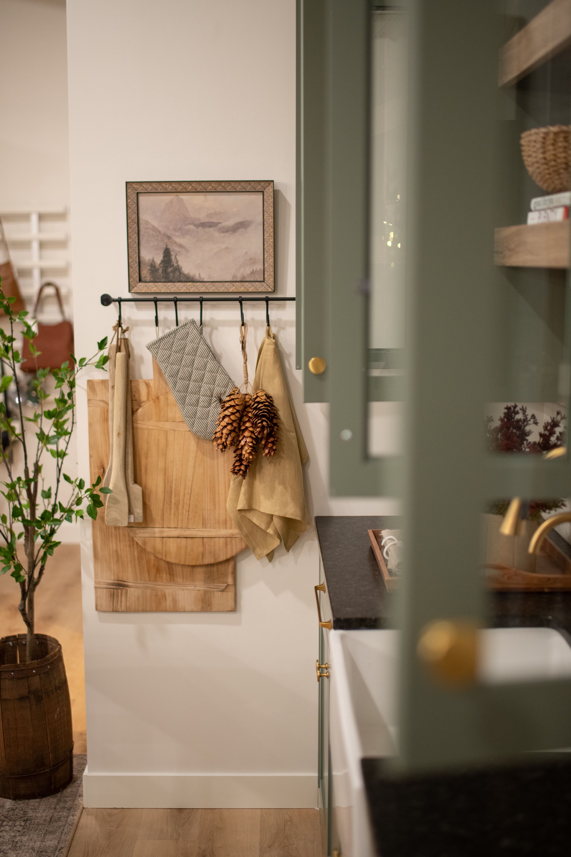 A brand photo of a cozy rustic kitchen corner with wooden cutting boards, pine cones, and framed artwork on sage green wall in a small shop.