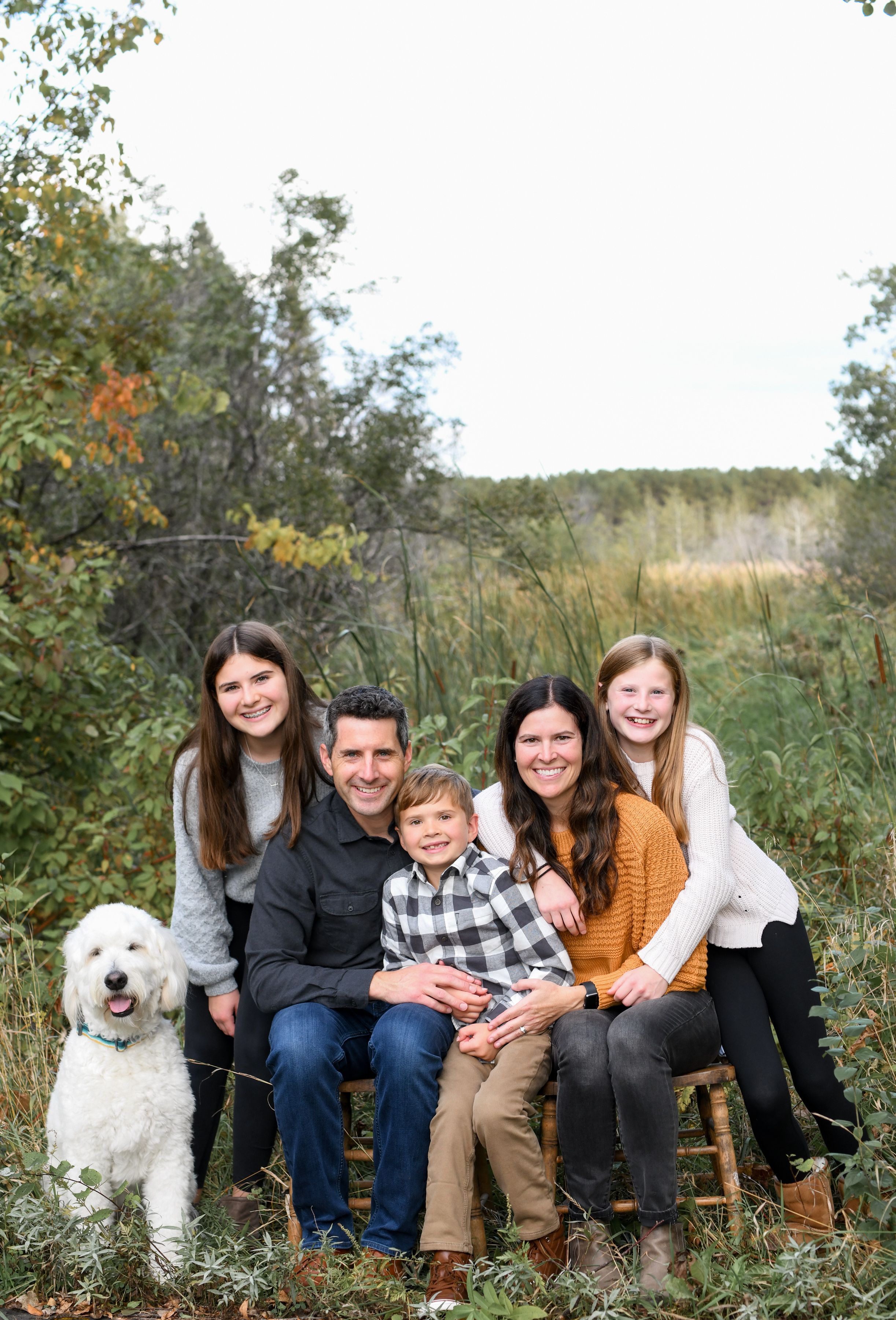 Portrait of a cozy family with a white dog sitting together outdoors, snuggling in a natural setting with tall grass and trees.